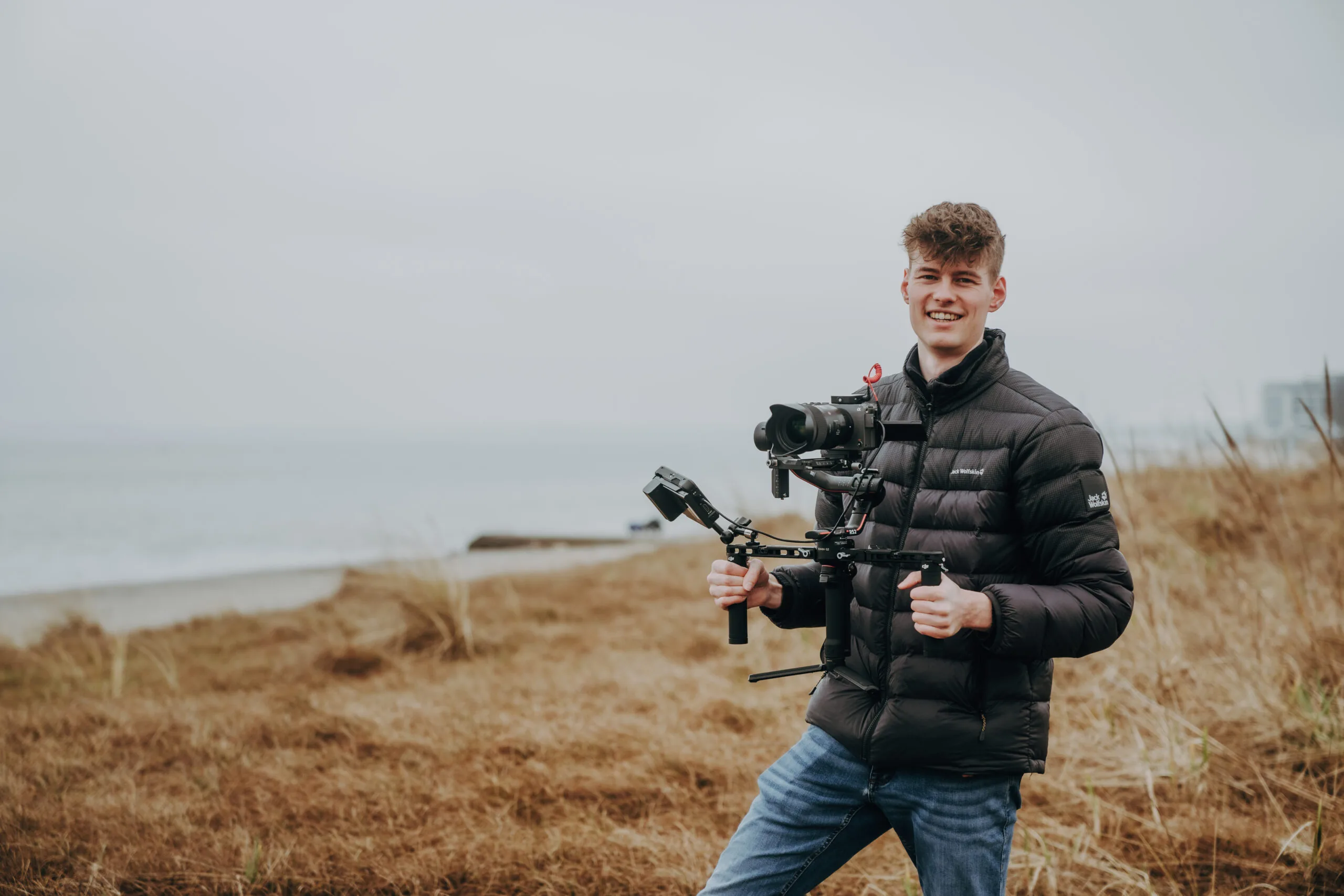 Junger Mann mit professioneller Kameraausrüstung und Gimbal lächelt in die Kamera, im Hintergrund ist eine Küstenlandschaft zu sehen.