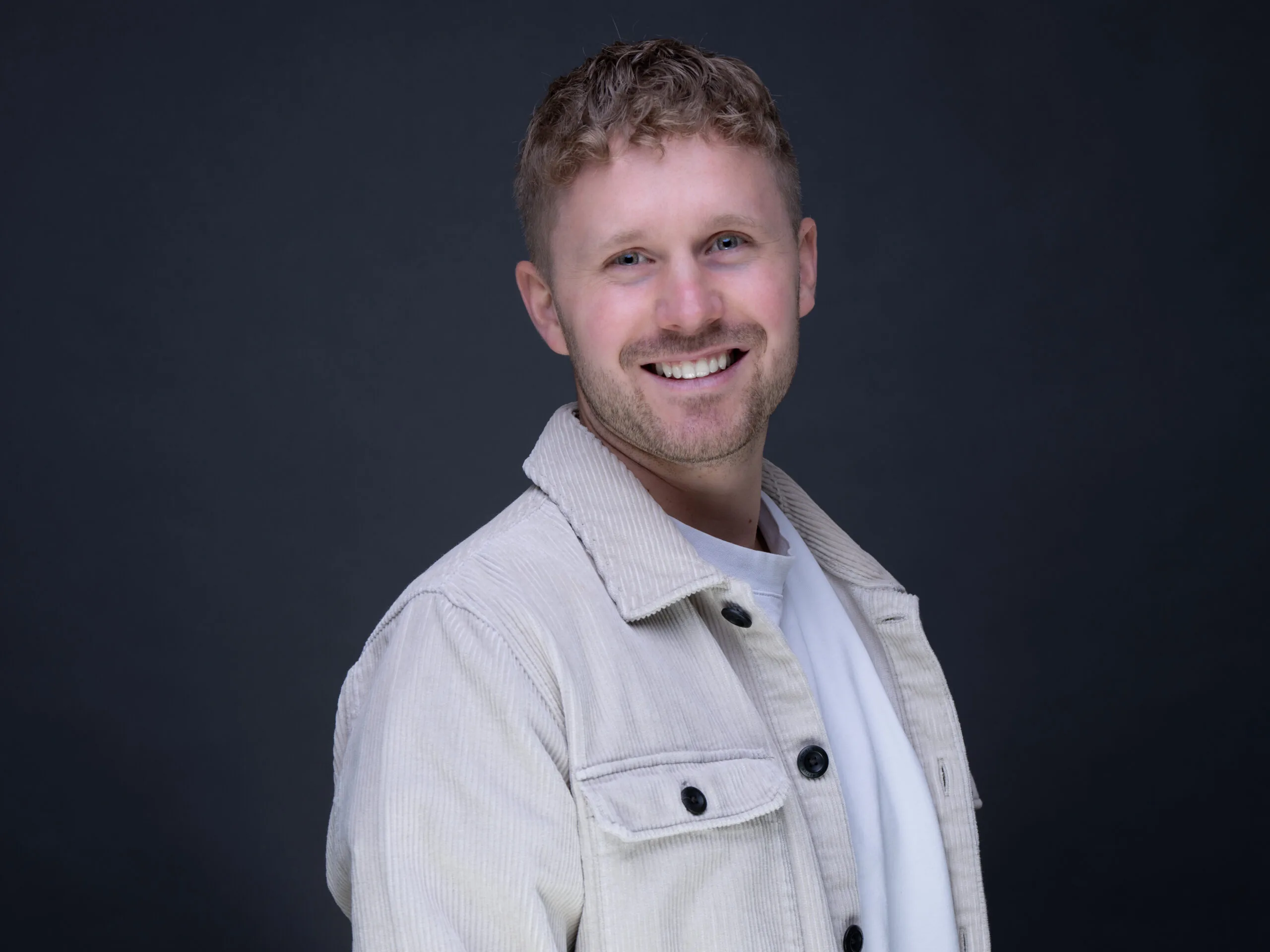 Ein lächelnder junger Mann mit blonden Locken und blauen Augen trägt eine helle Cordjacke vor dunklem Hintergrund.