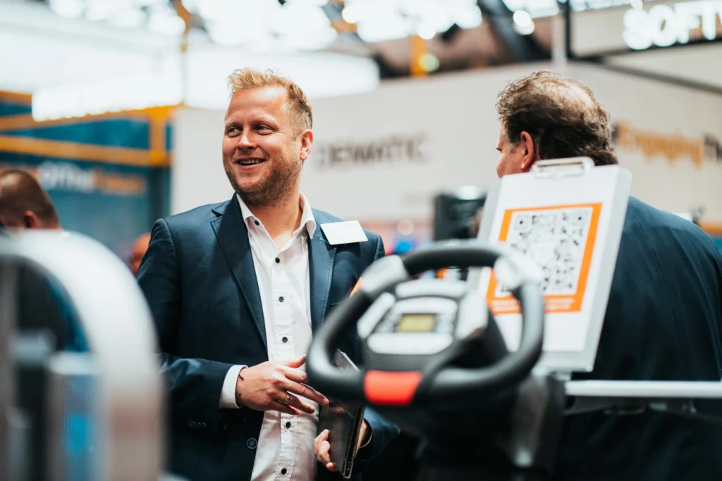 Ein lächelnder Mann in einem Anzug spricht auf einer Messe mit einer anderen Person, im Hintergrund sind weitere Stände zu sehen.