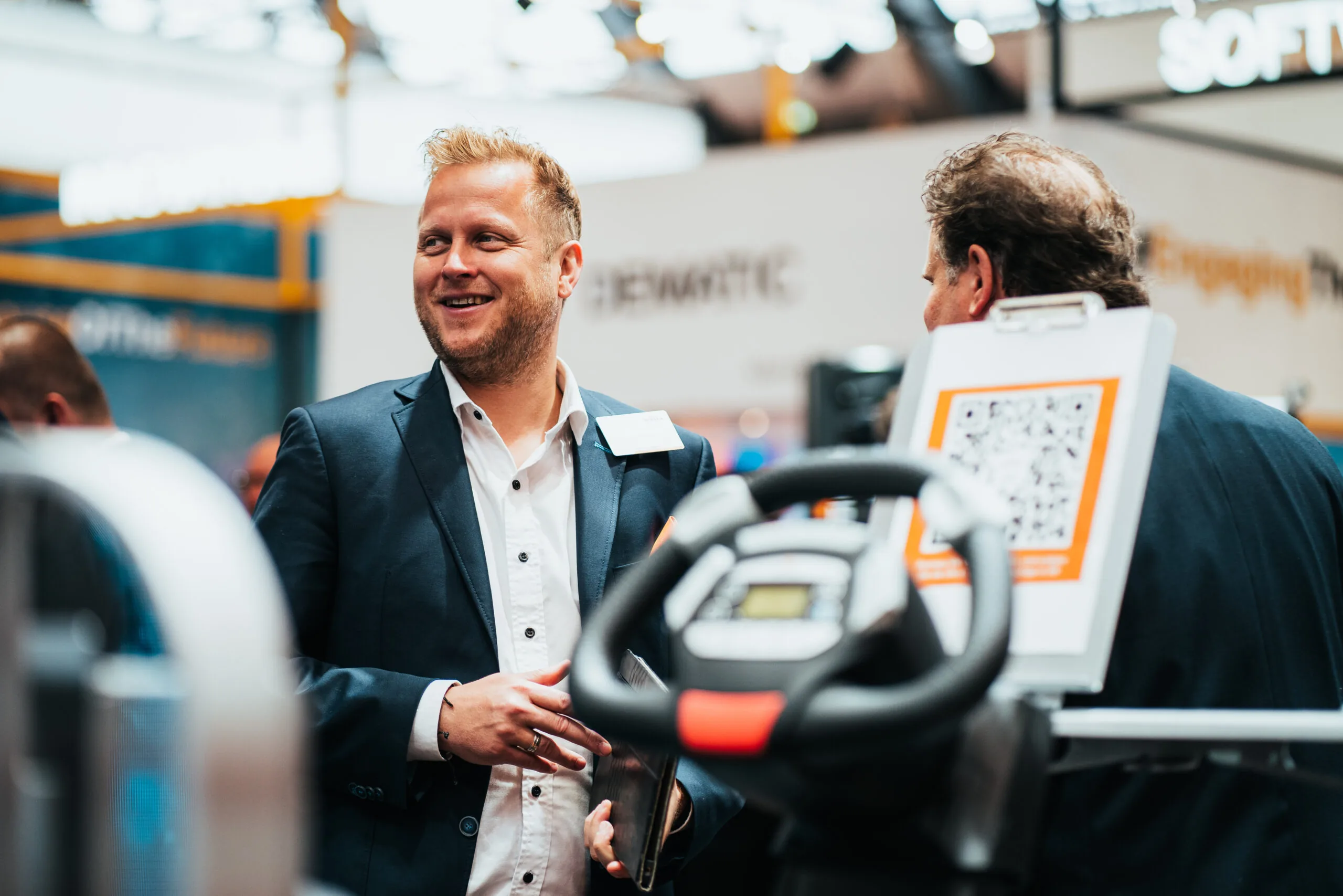 Ein lächelnder Mann in einem Anzug spricht auf einer Messe mit einer anderen Person, im Hintergrund sind weitere Stände zu sehen.