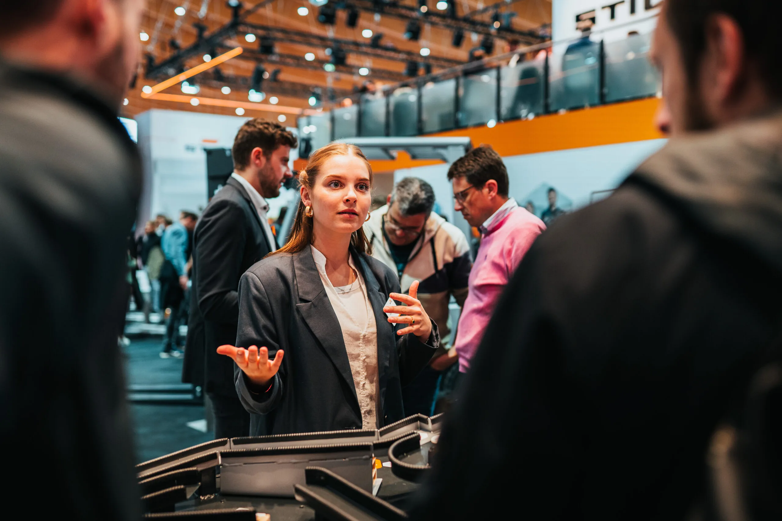 Eine junge Frau erklärt an einem Messestand ein technisches Modell, umgeben von weiteren Besuchern.