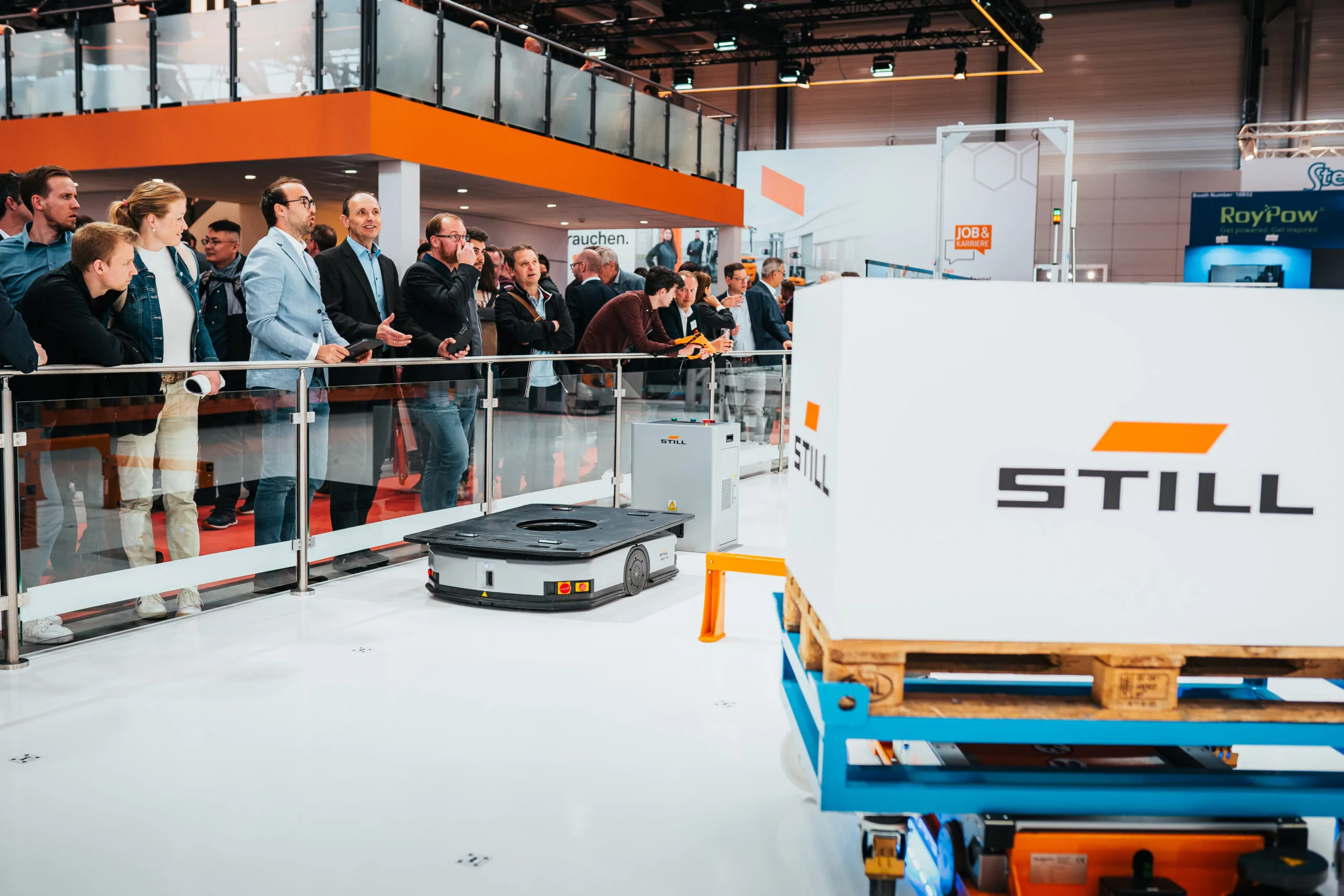 Besucher beobachten auf einer Messe ein automatisiertes Transportsystem und eine Palette mit dem STILL-Logo.