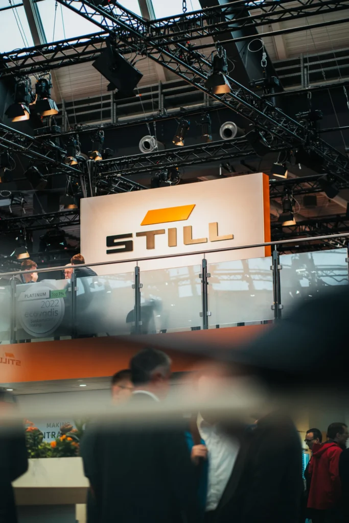 Das STILL-Logo auf einem großen Schild in einer Messehalle, umgeben von Beleuchtungstechnik und Besuchern.
