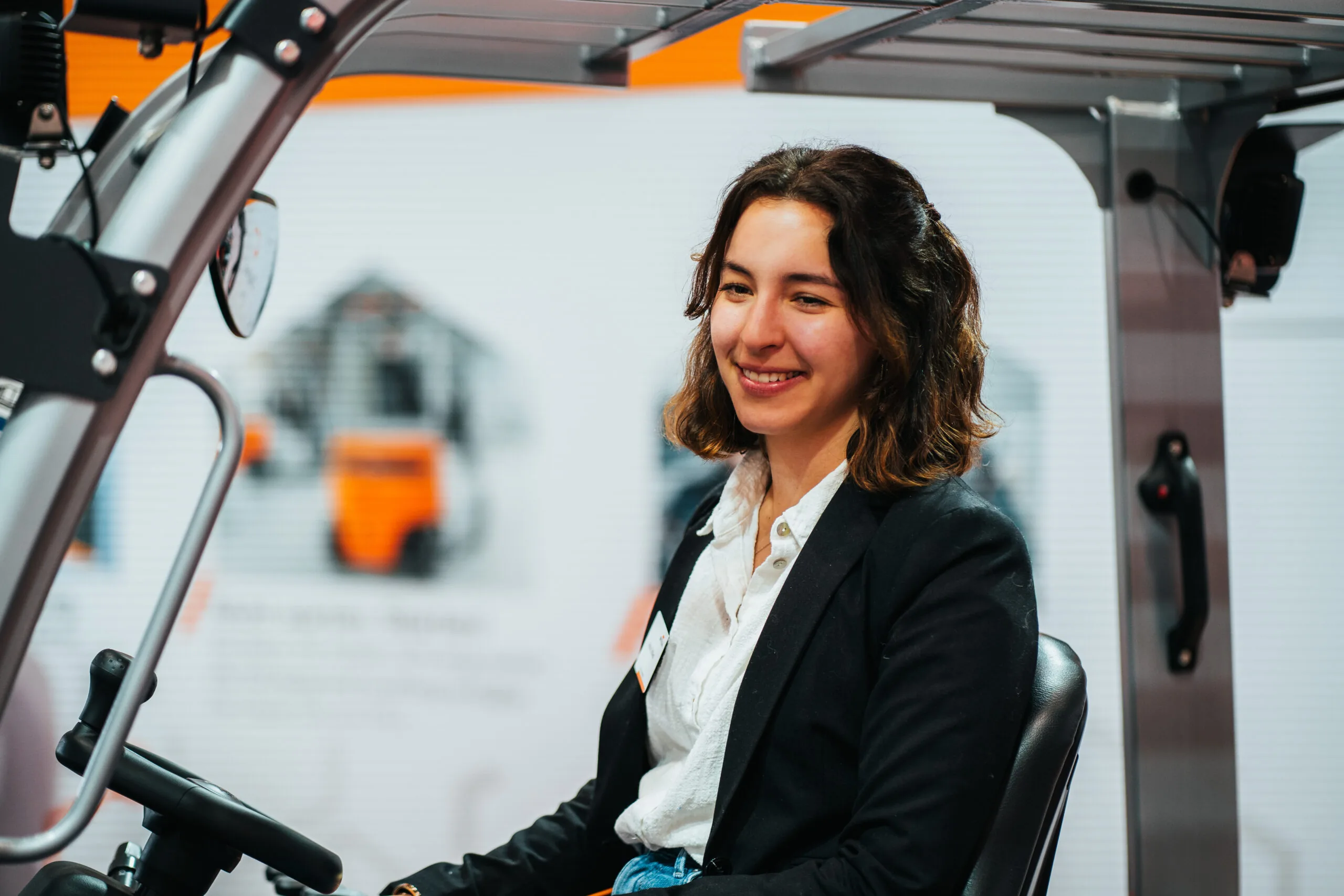 Eine lächelnde junge Frau sitzt am Steuer eines Fahrzeugs, möglicherweise eines Gabelstaplers, in einer hellen Industrieumgebung.