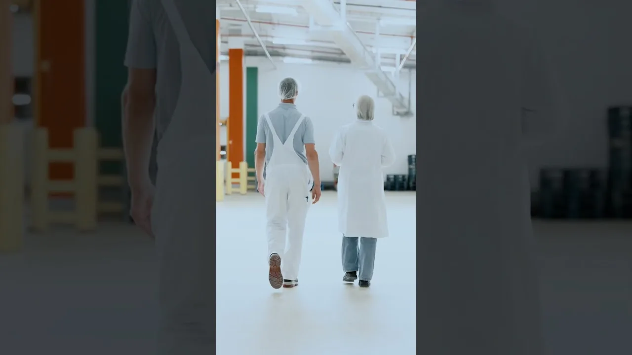 Zwei Personen in Schutzkleidung gehen durch eine saubere, hell beleuchtete Industrieanlage.
