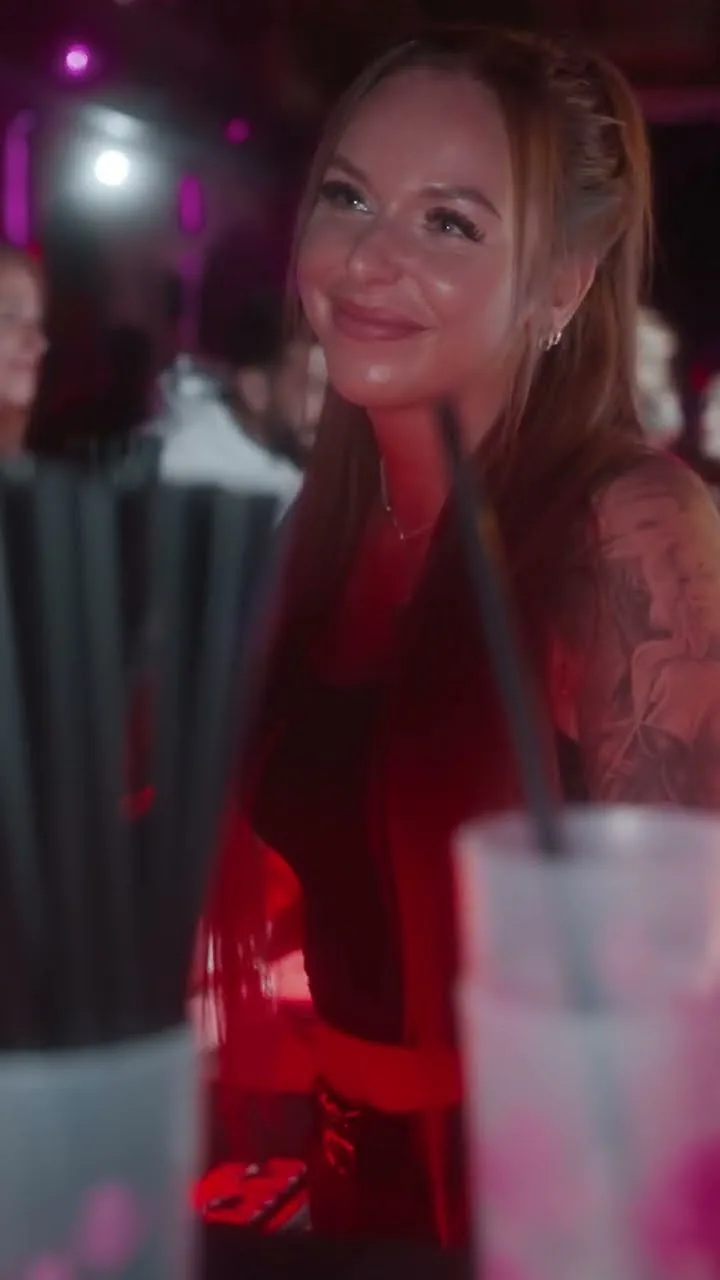 Eine lächelnde junge Frau mit langen, rötlich-braunen Haaren und einem Tattoo am Arm in einer lebhaften Bar- oder Clubumgebung.
