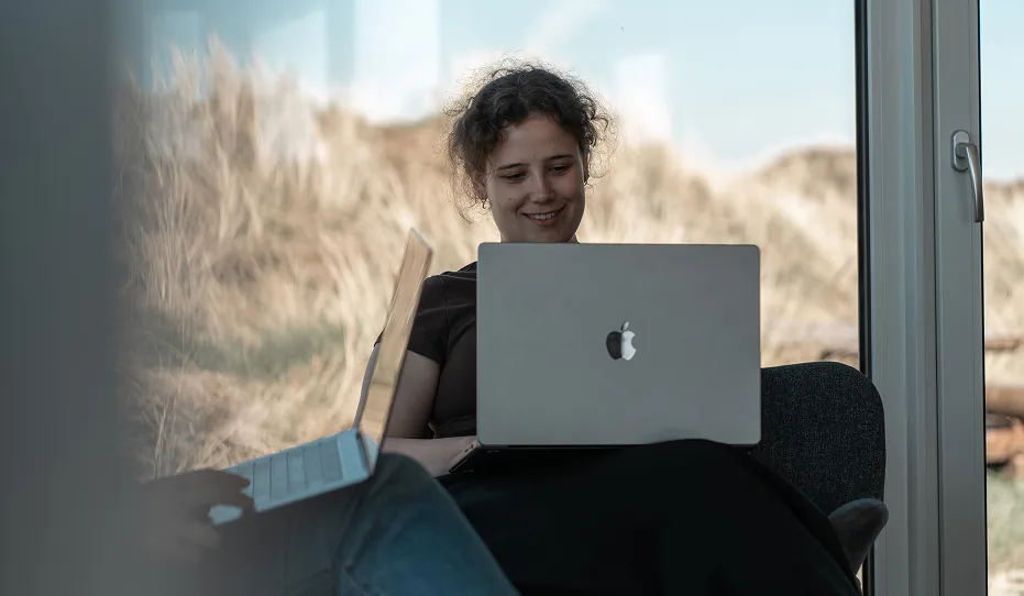 Eine lächelnde junge Frau mit lockigem Haar arbeitet an einem MacBook, während ein weiteres Laptop daneben steht, mit einer Dünenlandschaft im Hintergrund.