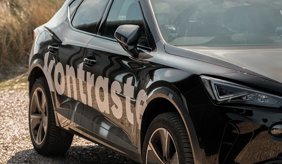 Ein schwarzes Auto mit dem Schriftzug 'kontraste' auf der Seite, geparkt auf einem Schotterweg mit trockenem Gras im Hintergrund.