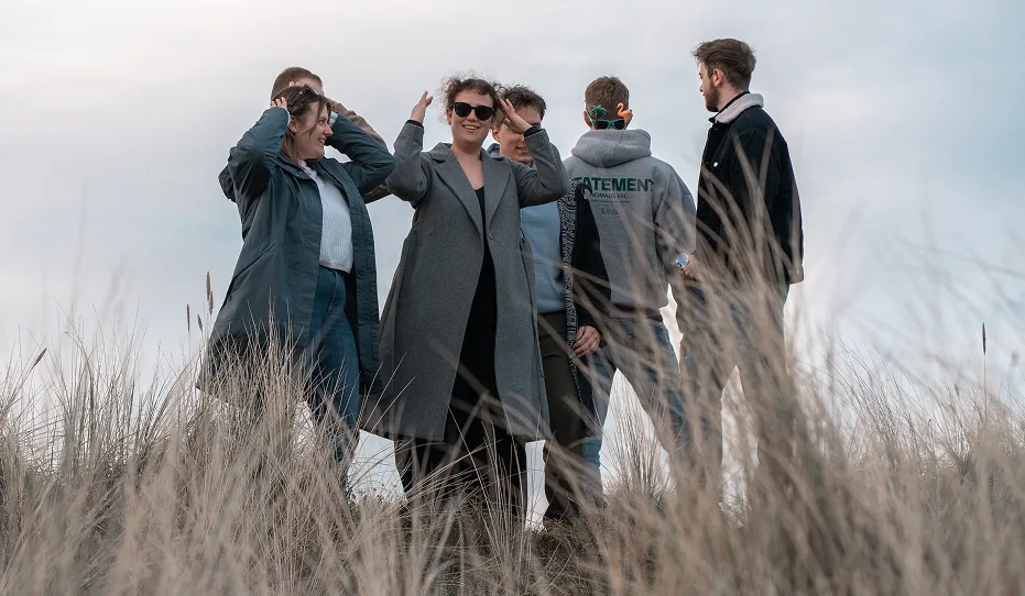 Ein junges Team steht lachend in hohem Gras unter einem bewölkten Himmel, die zentrale Person trägt Sonnenbrille und lächelt.
