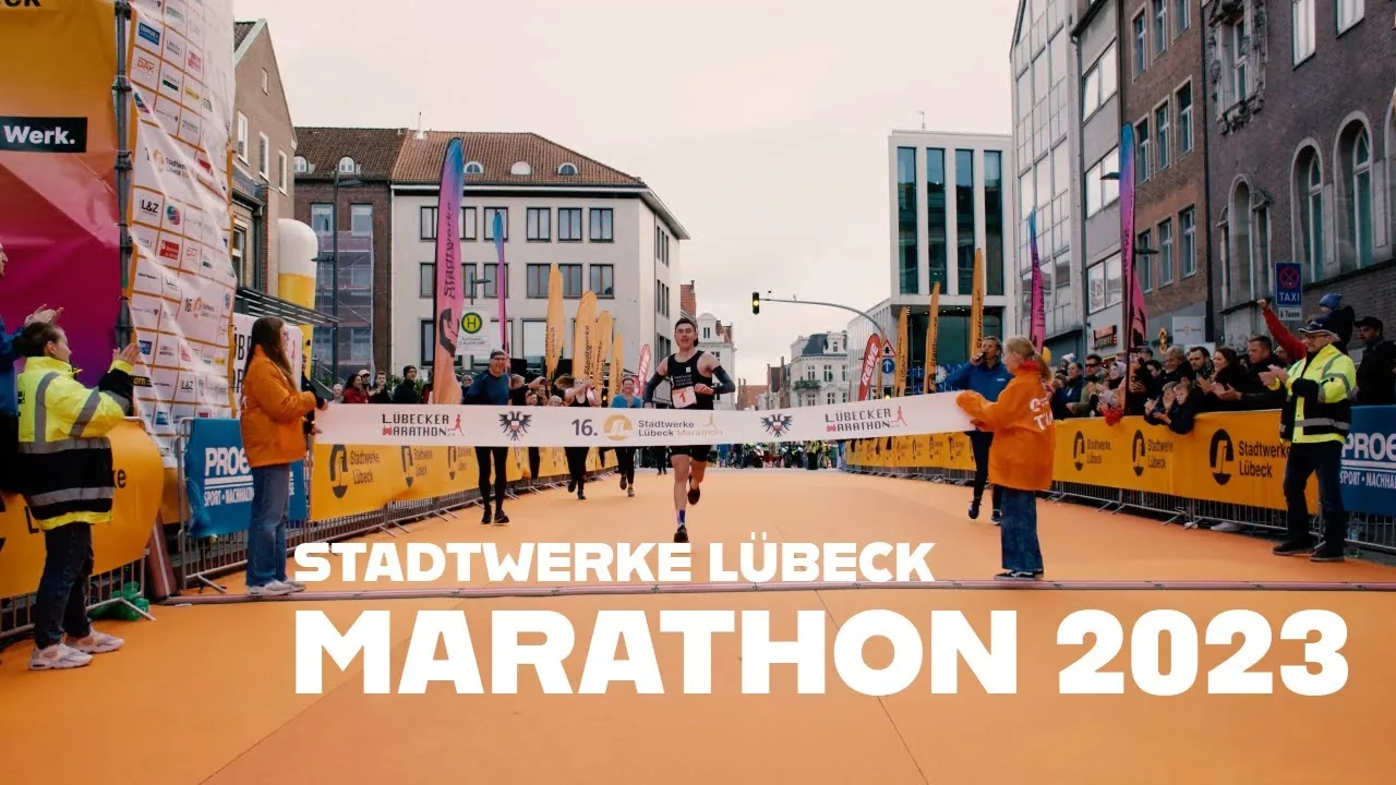 Ein Läufer überquert die Ziellinie des Stadtwerke Lübeck Marathons 2023, umgeben von jubelnden Zuschauern und Eventpersonal.