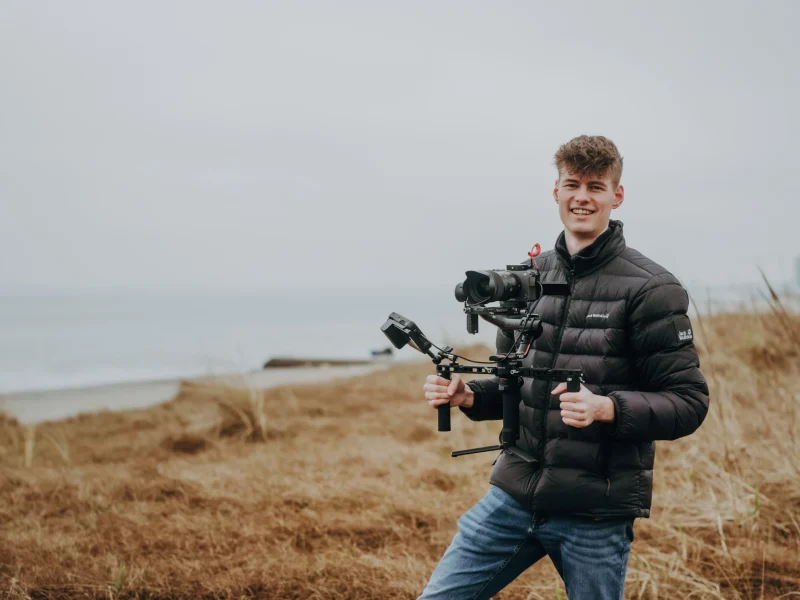 Junger Mann mit professioneller Kameraausrüstung und Gimbal lächelt in die Kamera, im Hintergrund ist eine Küstenlandschaft zu sehen.